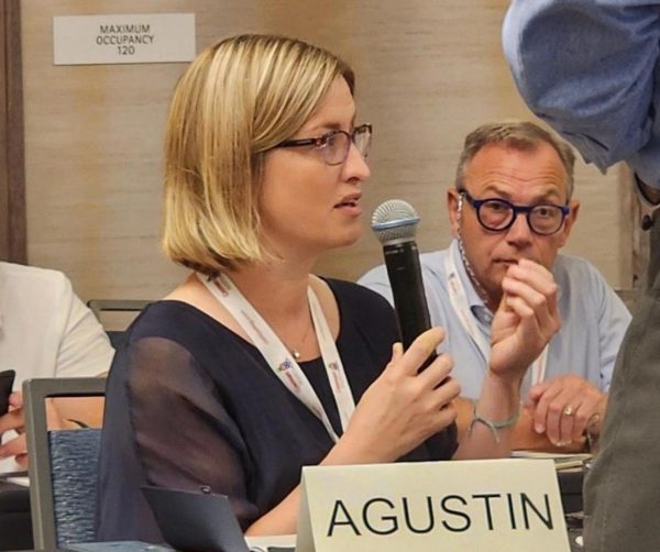 A woman with blonde hair and glasses speaks into a microphone at a conference on AI Governance & Risk Management, holding it in her right hand. She wears a lanyard and sits behind a nameplate labelled AGUSTIN, surrounded by other attendees.
